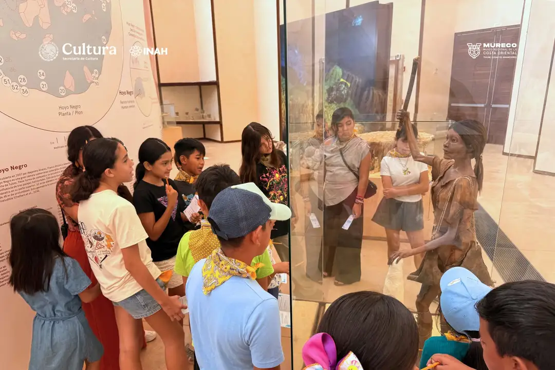 A group of children stand in a museum gallery viewing exhibition displays and listening to a guide explain artifacts and information panels