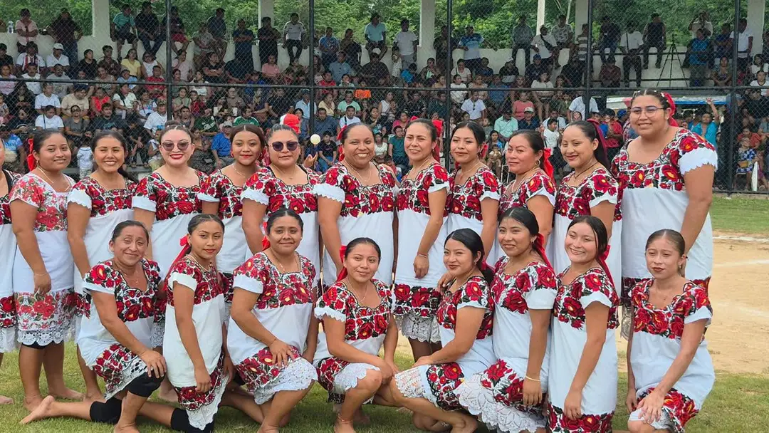 A women's softball team in matching white and red embroidered uniforms poses on a baseball field with spectators in the stands behind them