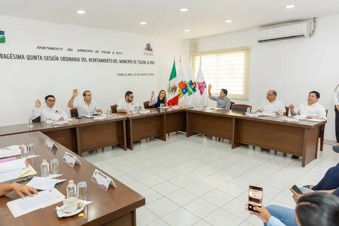 Municipal officials and attendees sit around wooden tables during a government meeting in an official chamber with Mexican flags displayed on the wall