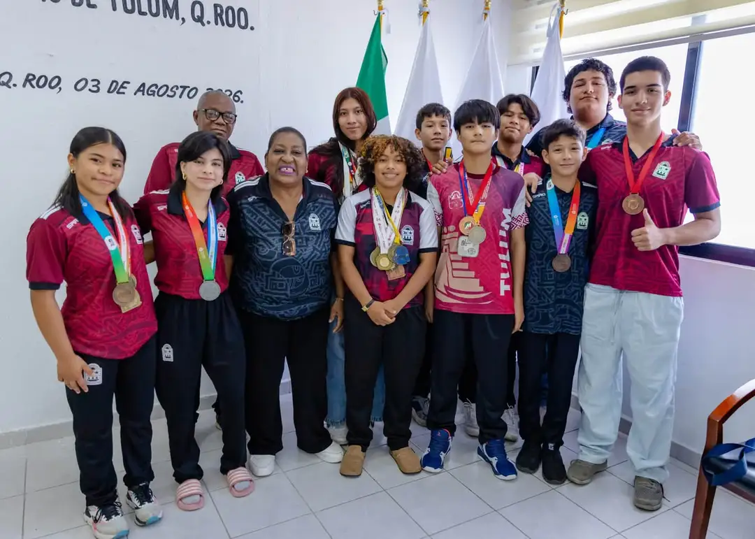Group of young athletes wearing maroon and dark uniforms with medals stand together in front of flags and officials indoors