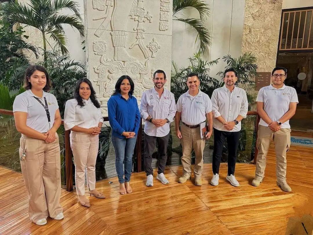 Seven people stand in a line on a wooden floor in front of a decorative stone wall and potted palms indoors