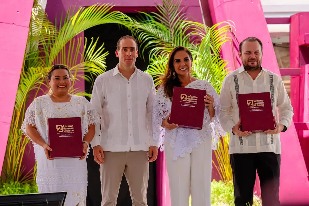 Four people in white clothing stand holding maroon books in front of bright pink architectural features and yellow-green palm fronds