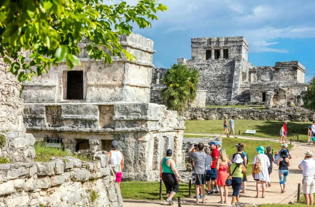 Ancient Walls, Caribbean Cliffs, and a Practical Guide to the Tulum Ruins Tourists walk among stone ruins with carved structures in a grassy courtyard beneath a blue sky