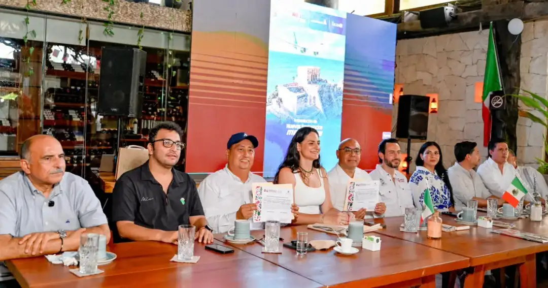 Nine people sit at a long wooden table during what appears to be a press conference or public announcement event, with colorful banners and a flag visible behin