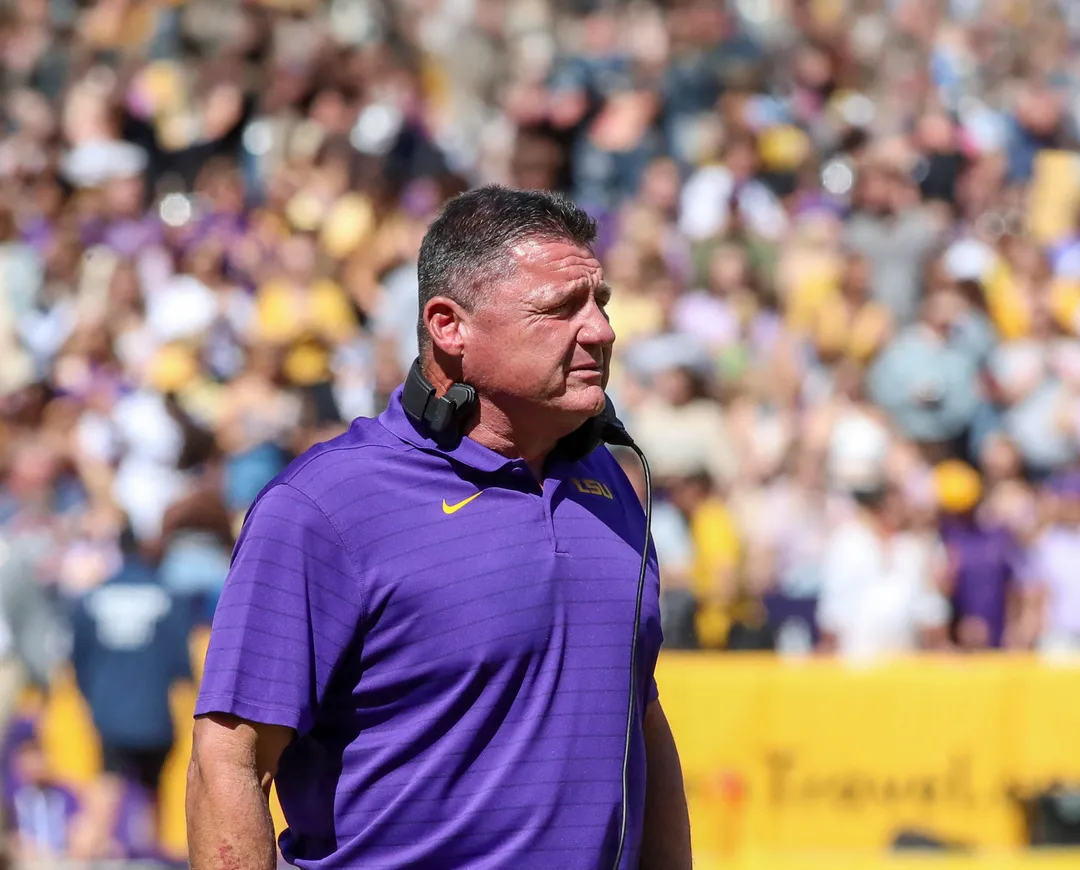 Man in purple polo shirt stands on sideline with crowded stadium in background