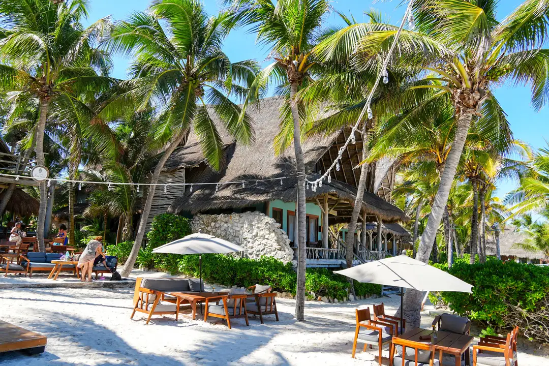 Ahau Tulum and the Quiet Intelligence of Hospitality Beachfront resort with thatched-roof bungalows surrounded by palm trees, white umbrellas, and wooden furniture on sand under clear blue sky