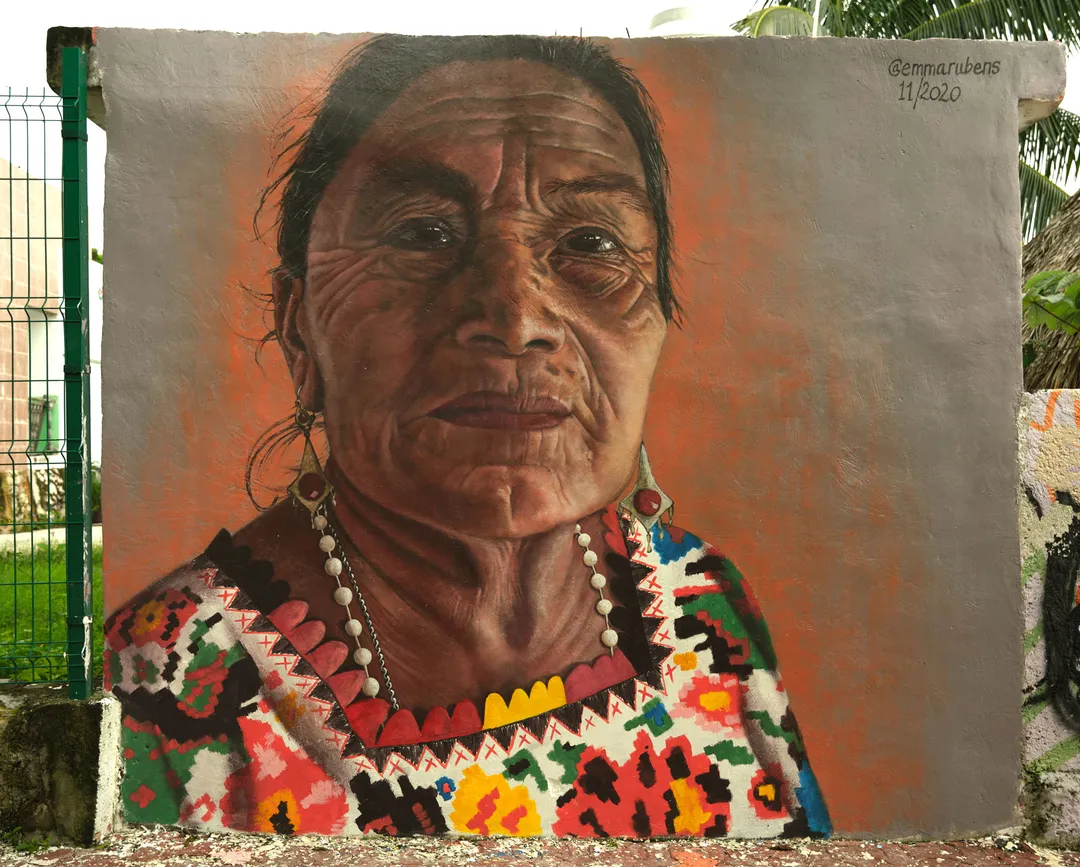 Mural portrait of an older woman wearing traditional colorful geometric patterned clothing and beaded necklace against an orange background