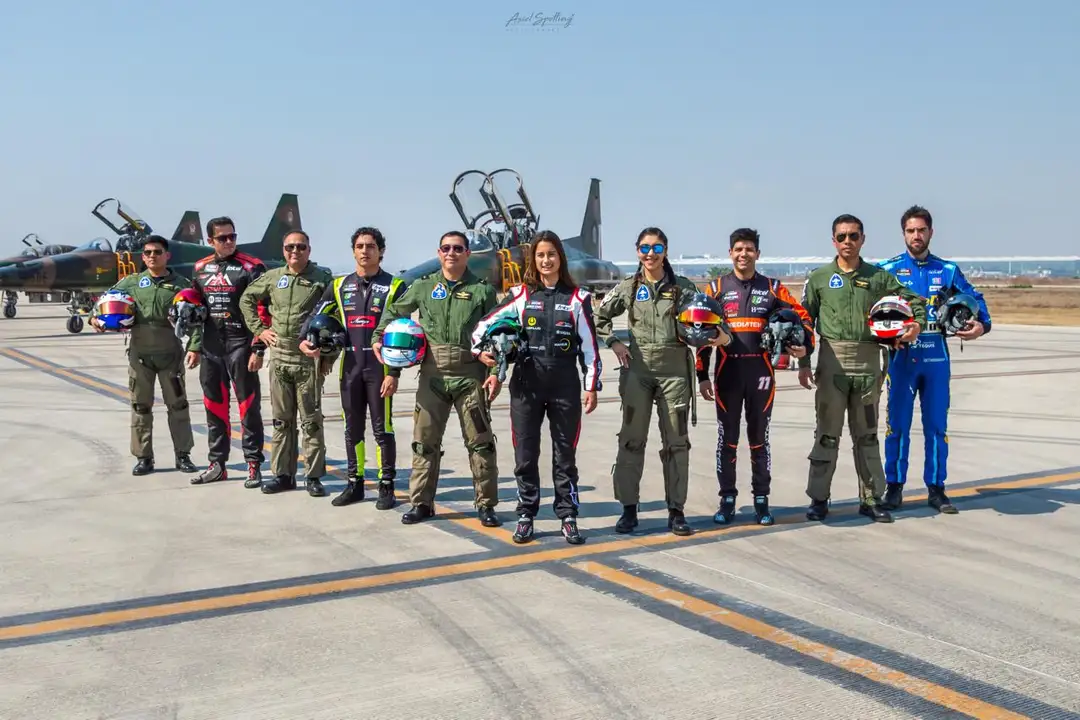 Group of pilots and aviation personnel in flight suits standing on an airport tarmac with a military jet aircraft in the background