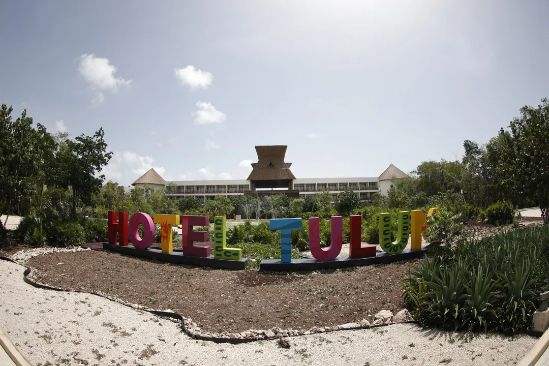 Colorful "Hotel Tulum" letters stand in a garden area with a large building and landscape visible behind them under a blue sky with white clouds