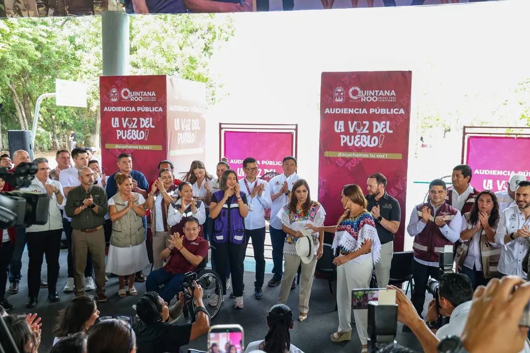 Tulum residents get direct access to state finance services A crowd of people gathers at an outdoor public event with burgundy banners reading "Audiencia Pública" and "La Voz del Pueblo," with attendees applauding and re
