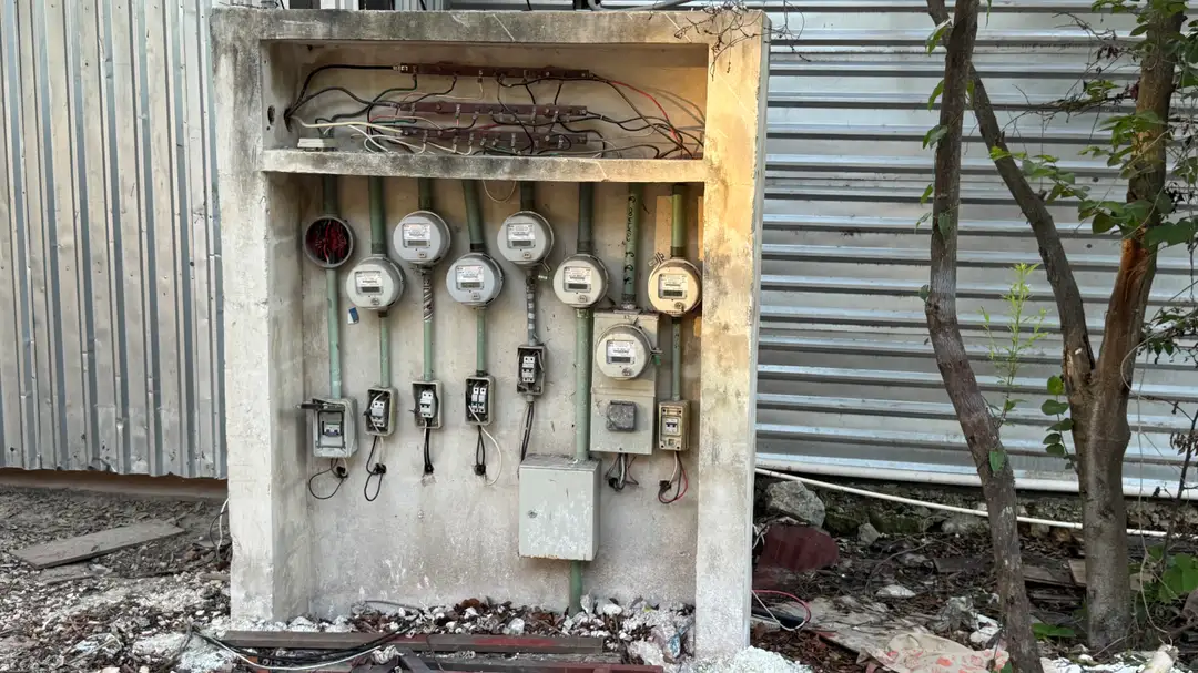 Anyone Can Touch These Live Wires, and in Tulum Nobody Is Checking Multiple utility meters and electrical switches mounted on a weathered concrete box beside metal structures and a small tree