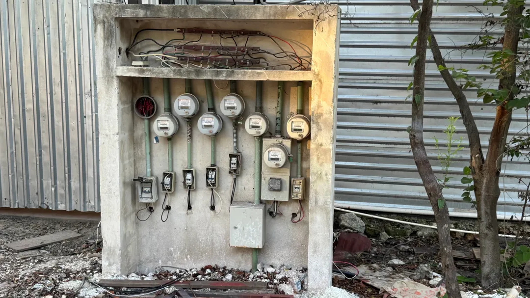 Multiple utility meters and electrical switches mounted on a weathered concrete box beside metal structures and a small tree