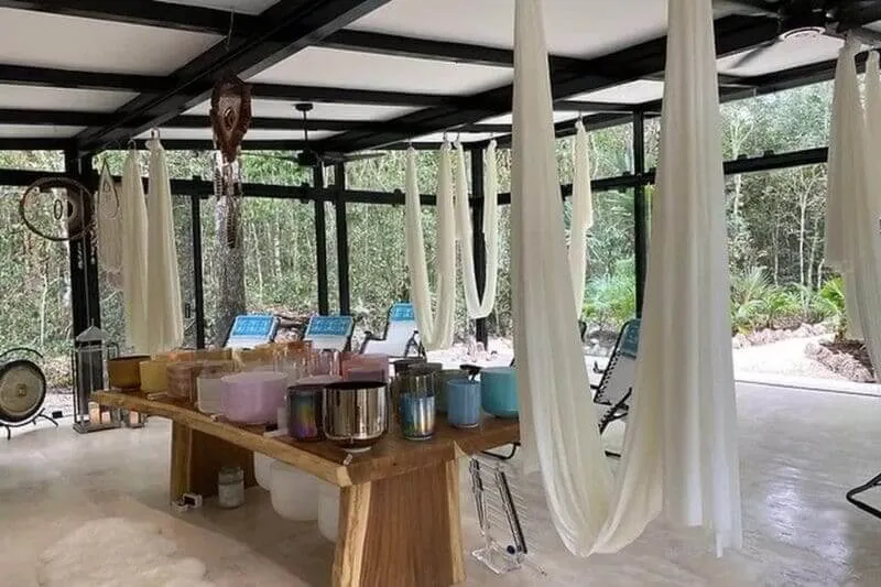 Sound Healing Tulum: A Gateway to Authentic Healing and Self-Discovery A wooden table with metal bowls and containers sits in a covered pavilion with flowing white curtains, forest visible through large windows