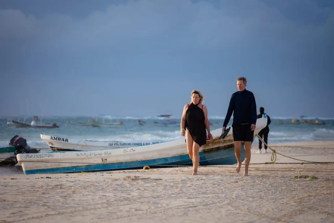 Surfers in wetsuits walk along a sandy beach past moored fishing boats under clear skies