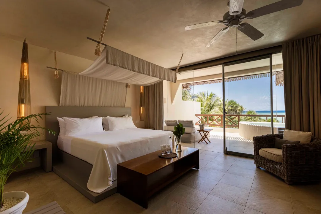 Jashita Hotel Affiliates With Relais & Châteaux, Redefining Luxury in Tulum Upscale hotel bedroom with canopy bed, ocean view through glass doors, and tropical landscape visible beyond the balcony