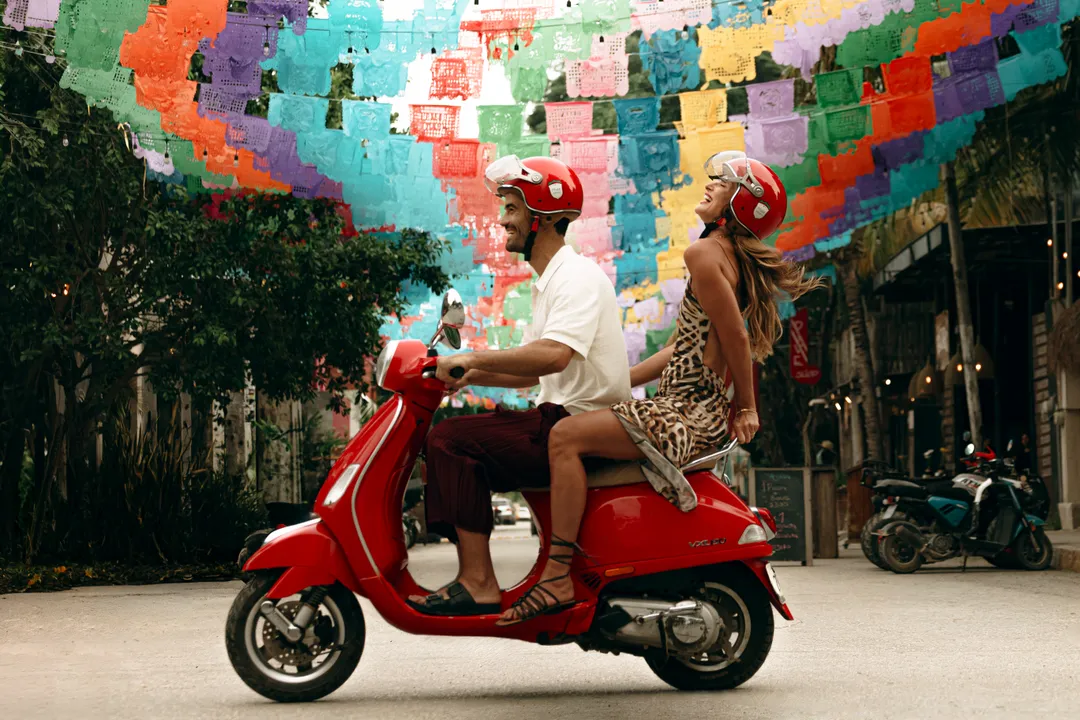 Red against Caribbean blue, the campaign that photographs Tulum as one single world Two people wearing red helmets ride a red scooter down a narrow street beneath a colorful overhead canopy of paper flags
