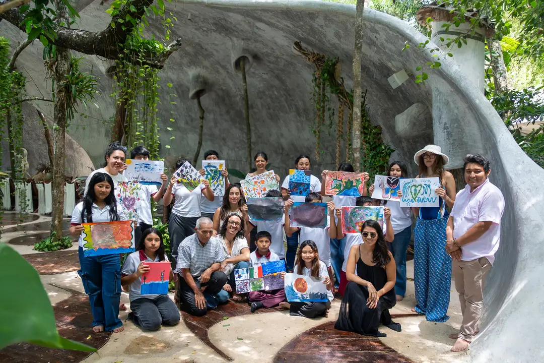 Letras Itinerantes Brings Manuel Antonio Ay Youth to One of the Riviera Maya's Top Art Spaces A group of young people and adults display colorful artwork and drawings in front of a large curved concrete wall covered with vines and climbing plants