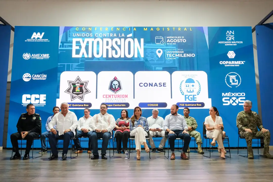 A group of officials and representatives sit on a stage in front of a blue banner advertising a magistrate conference on extortion, with sponsor logos displayed