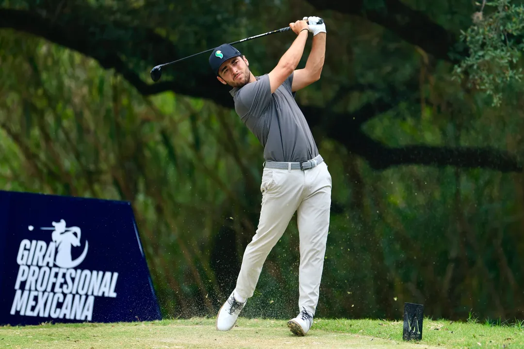 Golfer in gray shirt and cap swings club on grass course with green trees and tournament signage in background