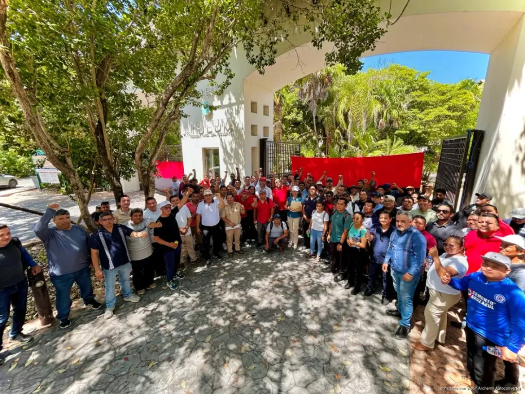 Kore Tulum Workers Strike Over Unpaid Benefits After Hotel Closes Without Warning A large group of people gather in a courtyard next to a white building with red banners, standing on a stone pathway shaded by trees