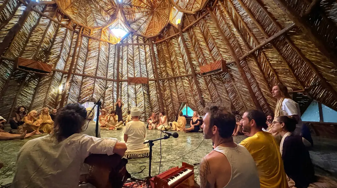 Barefoot in Azulik's Dome, Nearly 100 People Opened a Week of Yoga and Music People sit barefoot inside a large wooden dome structure with woven palm frond walls and an open ceiling, watching performers with musical instruments