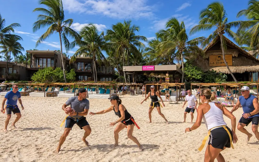 Bills Mafia Will Watch the Patriots Game From a Swim-Up Bar in Tulum Multiple people play volleyball on a sandy beach lined with palm trees and thatched-roof beach clubs under a clear blue sky