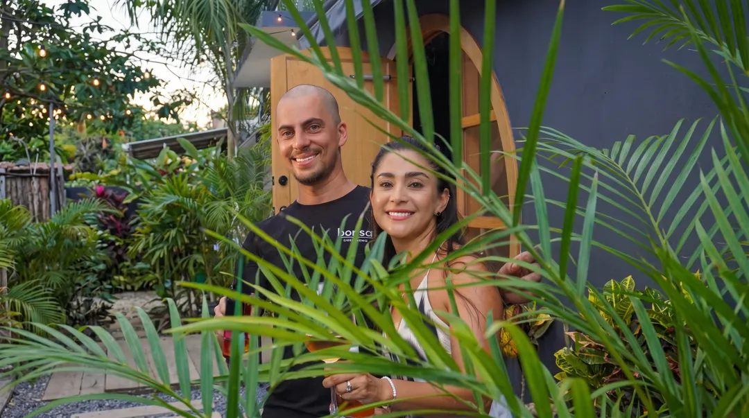 Dos personas posan entre macetas con plantas y palmas frente a un edificio de paredes amarillas y azul oscuro
