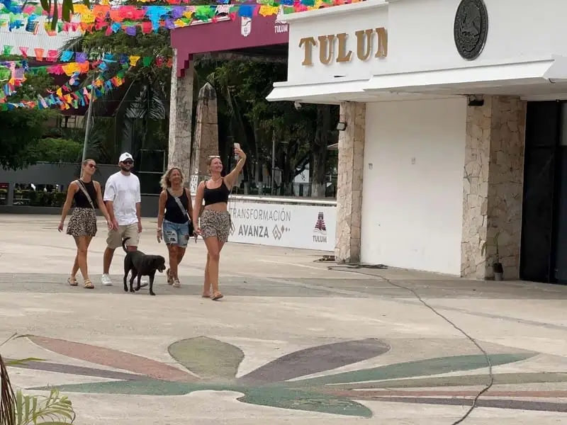 Four people and a small black dog walk past a white storefront with colorful paper decorations strung above in a town plaza