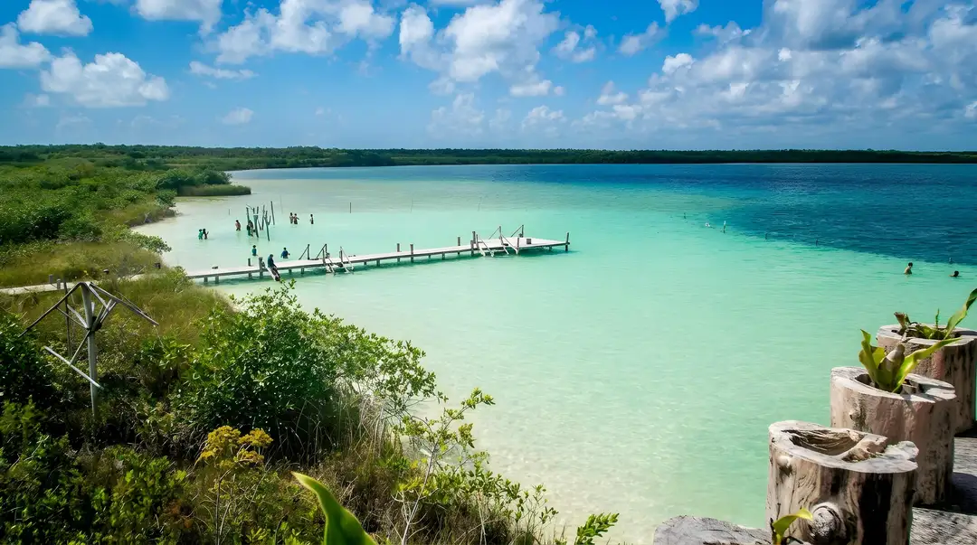 Wooden pier extends into shallow turquoise water lined by mangrove vegetation under blue sky with scattered clouds