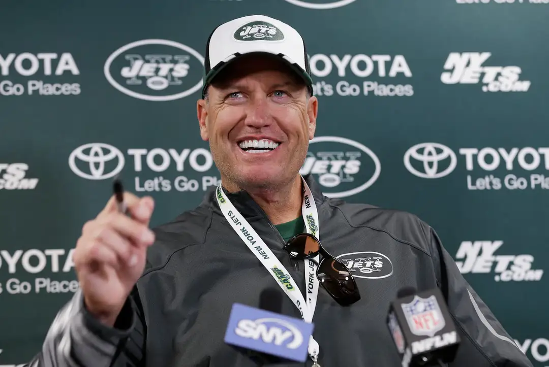 Man in Jets cap and jacket gestures while speaking at a press conference with team logos in the background