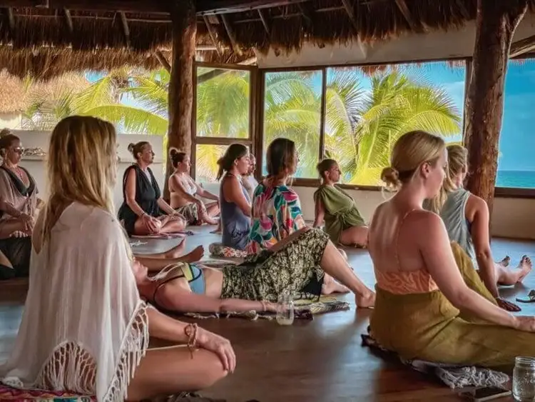 A group of people sit on the floor of a beachside pavilion with thatched roof and ocean views through large windows