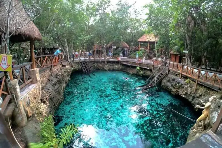 Turquoise cenote surrounded by wooden walkways and thatched-roof structures with visitors exploring the natural sinkhole Turquoise cenote surrounded by wooden walkways and thatched-roof structures with visitors exploring the natural sinkhole