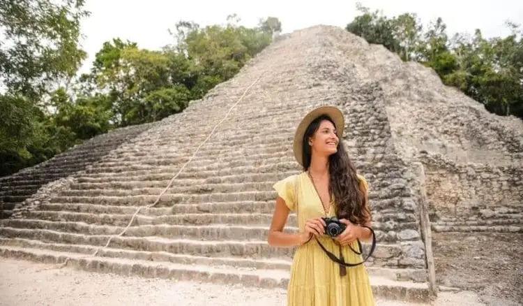 Woman in yellow dress and hat holds a camera while standing in front of a stepped stone pyramid surrounded by trees