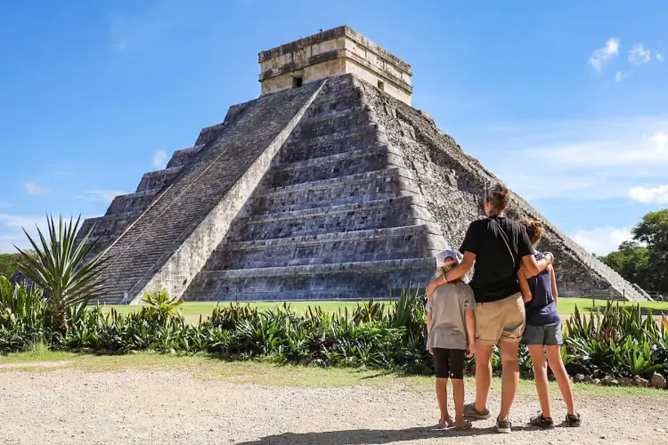 Three visitors stand on a path viewing a large stepped pyramid with a temple structure on top, surrounded by vegetation and green grass