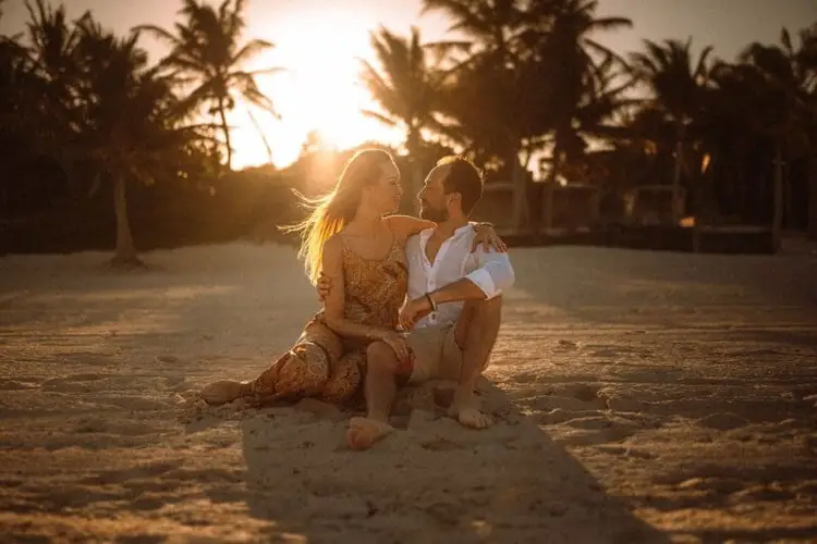 Two people sit together on sand at sunset with palm trees silhouetted in the background
