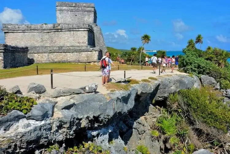 A visitor walks through an ancient stone structure overlooking the ocean, with tourists and palm trees visible at the archaeological site