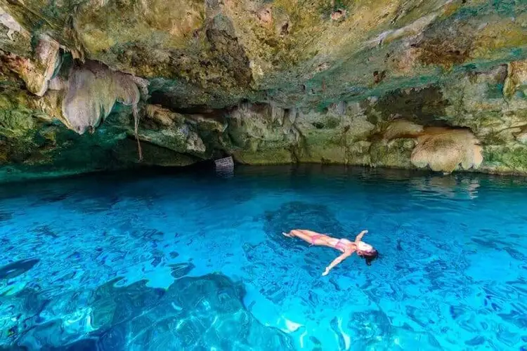 Person floating in bright blue water inside a cave with moss-covered rock formations and stalactites overhead