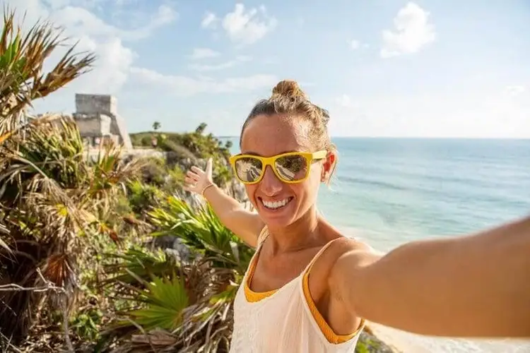 Woman wearing yellow sunglasses takes a selfie on a coastal cliff with tropical vegetation and a stone structure visible behind her overlooking the ocean Woman wearing yellow sunglasses takes a selfie on a coastal cliff with tropical vegetation and a stone structure visible behind her overlooking the ocean