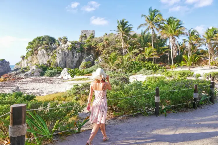 Woman in a pink dress and white hat walking on a sandy path with palm trees and rocky cliffs in the background Woman in a pink dress and white hat walking on a sandy path with palm trees and rocky cliffs in the background