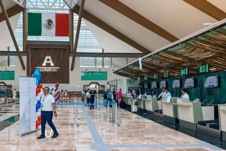 Airport terminal interior with Mexican flag overhead, check-in counters along the right wall, and travelers moving through the spacious hall with modern archite