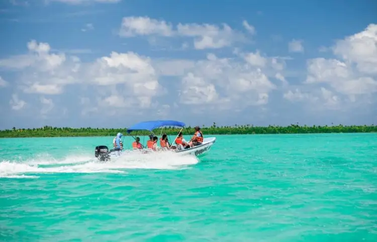 A motorboat with several passengers navigates turquoise water near a tropical shoreline under a partly cloudy sky