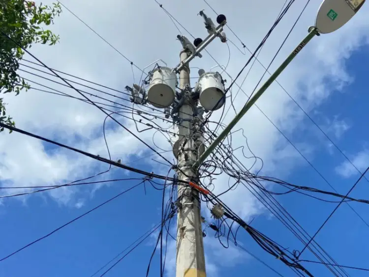 Utility pole with multiple tangled power lines, transformers, and street lights against a blue sky with clouds Utility pole with multiple tangled power lines, transformers, and street lights against a blue sky with clouds