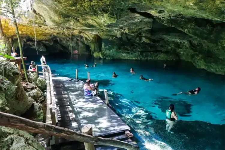 Swimmers and visitors enjoy a turquoise underground pool within a cave surrounded by moss-covered limestone rocks and wooden walkways