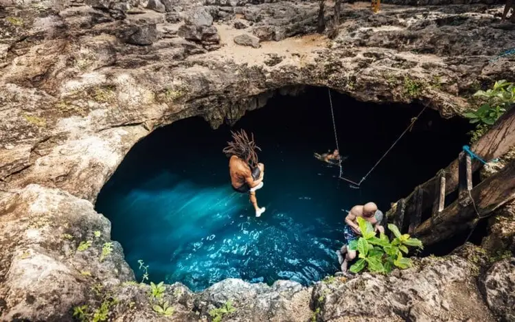 Person swinging on rope into a blue cenote surrounded by rocky limestone cliffs Person swinging on rope into a blue cenote surrounded by rocky limestone cliffs