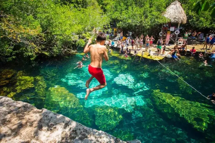 Shirtless man jumping into a clear turquoise cenote surrounded by dense jungle vegetation and visitors on a beach area with a thatched-roof structure