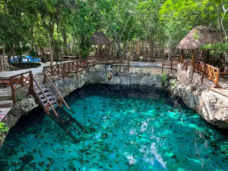 A turquoise cenote surrounded by limestone formations and wooden railings, with dense jungle canopy and thatched-roof structures in the background