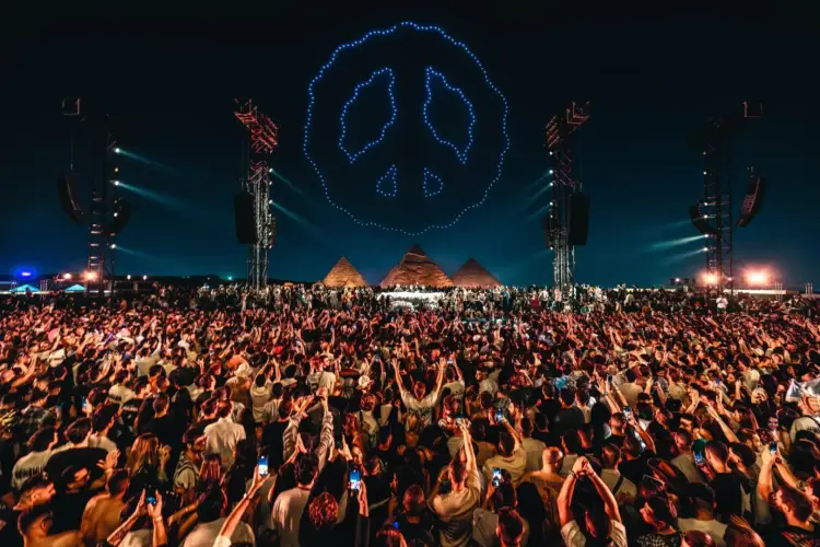 Large crowd with hands raised at a nighttime outdoor festival beneath a glowing blue peace symbol logo Large crowd with hands raised at a nighttime outdoor festival beneath a glowing blue peace symbol logo