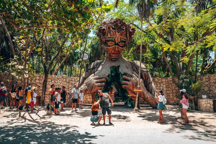 Visitors walk around a large wooden sculpture with a carved face in a tree-lined courtyard surrounded by stone walls