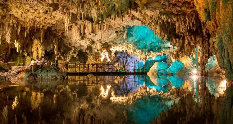 Underground cave with stalactites and a walkway crossing turquoise water that reflects the illuminated limestone formations above Underground cave with stalactites and a walkway crossing turquoise water that reflects the illuminated limestone formations above