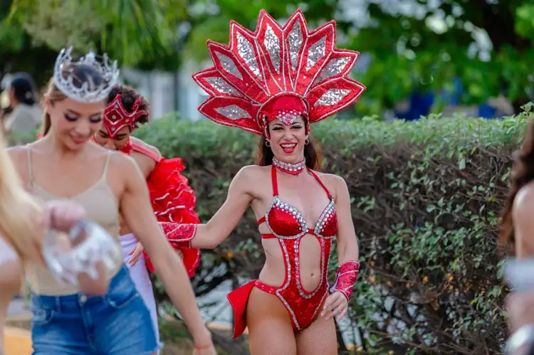 Performers in elaborate red and silver costumes with decorative headdresses parade outdoors past hedges during a carnival celebration